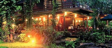 A traditional wooden building with a covered outdoor area, surrounded by lush greenery and a small waterfall.