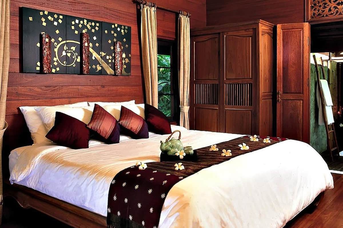 A wooden bed with white linens and dark pillows, a decorative wall piece, and a green teapot on the bedside table.