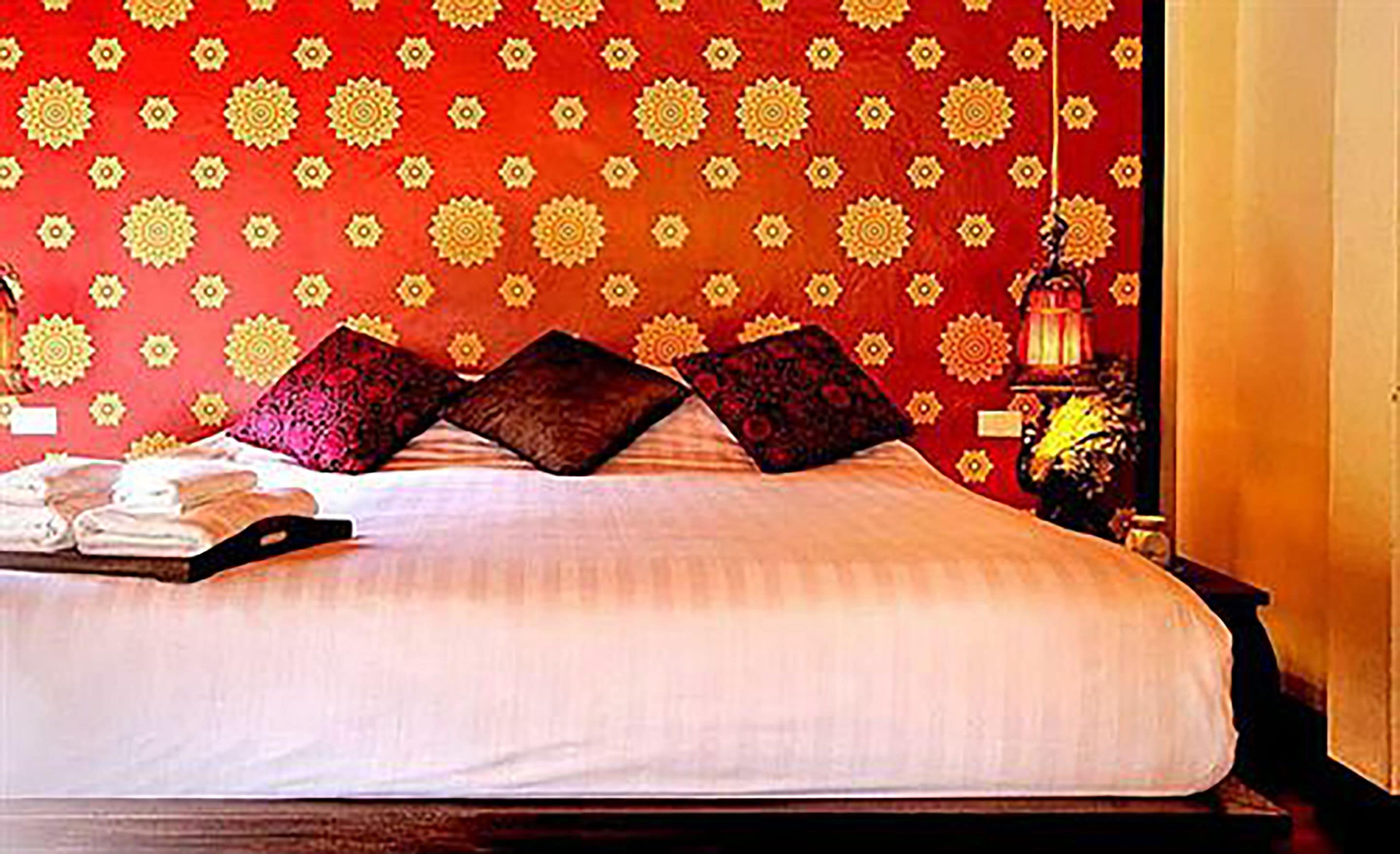 A neatly made bed with a white duvet, two dark pillows, and a red patterned wall in the background.