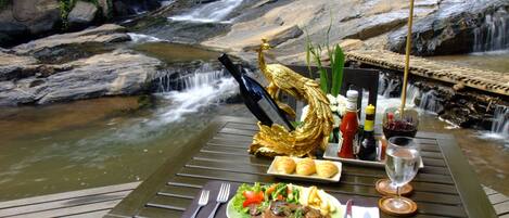 Une table à manger dressée avec un repas, une bouteille et un verre de vin rouge, le tout mis en valeur par un décor composé d’une cascade et de verdure.
