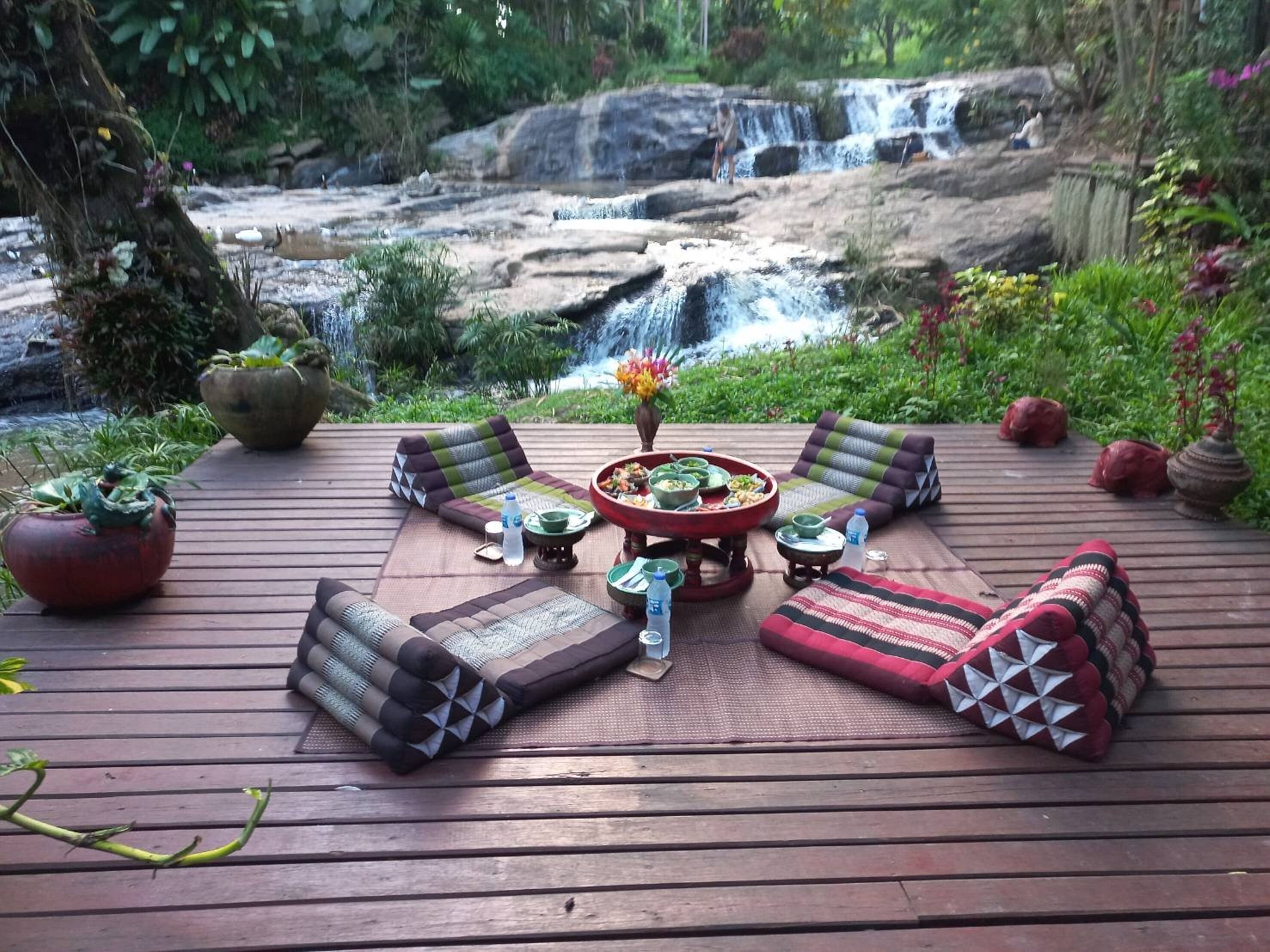 A wooden deck with outdoor seating, a central fire pit, and a waterfall in the background.