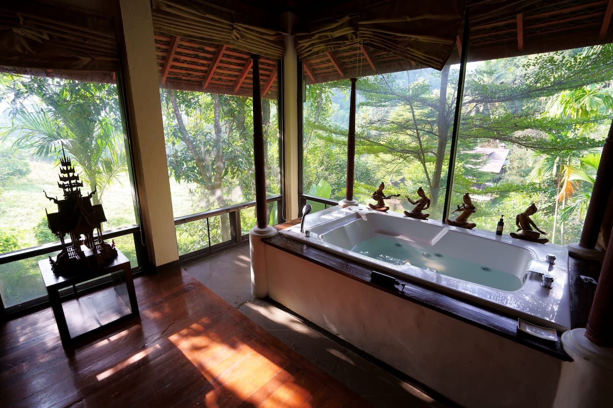 A room with a large bathtub, a small table with a decorative item, and a view of lush greenery through large windows.