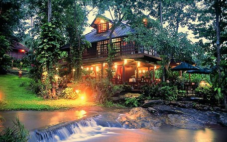 A large, two-story wooden house with a waterfall in front, surrounded by lush greenery.
