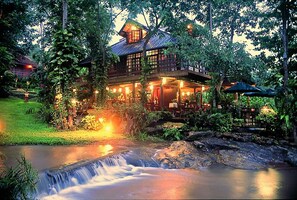 A large, two-story wooden house with a waterfall in front, surrounded by lush greenery. - Sukantara Cascade Resort & Spa (Mae Rim)