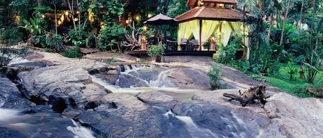 A waterfall flowing through a rocky landscape with a traditional wooden structure in the background.