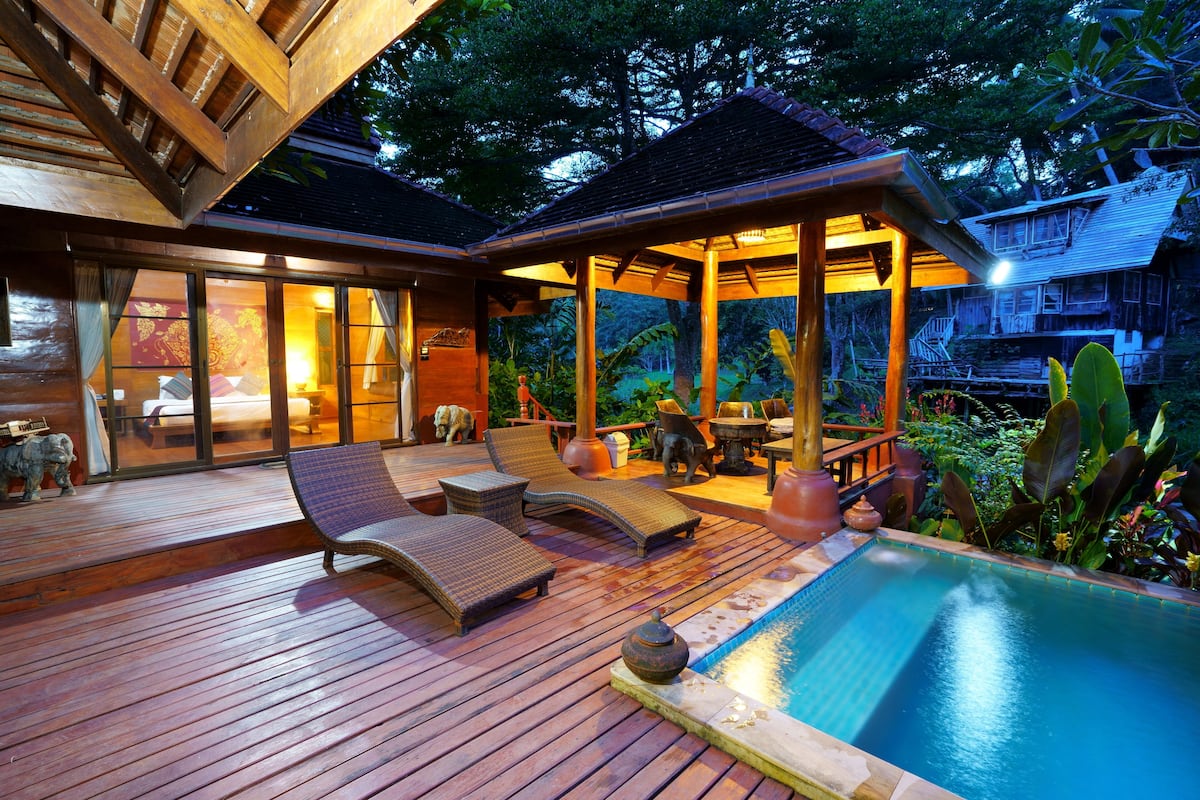 A wooden deck with lounge chairs and a swimming pool, surrounded by lush greenery and a wooden structure with a tiled roof.