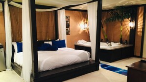 A four-poster bed with white bedding and blue pillows, a bedside table with a lamp, a small bathroom with a sink and mirror, and a blue round ottoman. - Hotel - Motel Coconut (Trois-Rivieres)