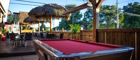 A pool table with a red felt surface, surrounded by outdoor seating and a thatched roof structure.