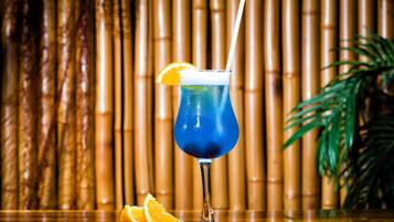 A blue cocktail with an orange slice garnish and a straw, placed on a wooden surface with a bamboo background.