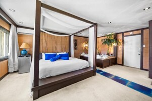 A four-poster bed with a canopy, a bedside table with a lamp, a small desk with a chair, and a door in a hotel room.