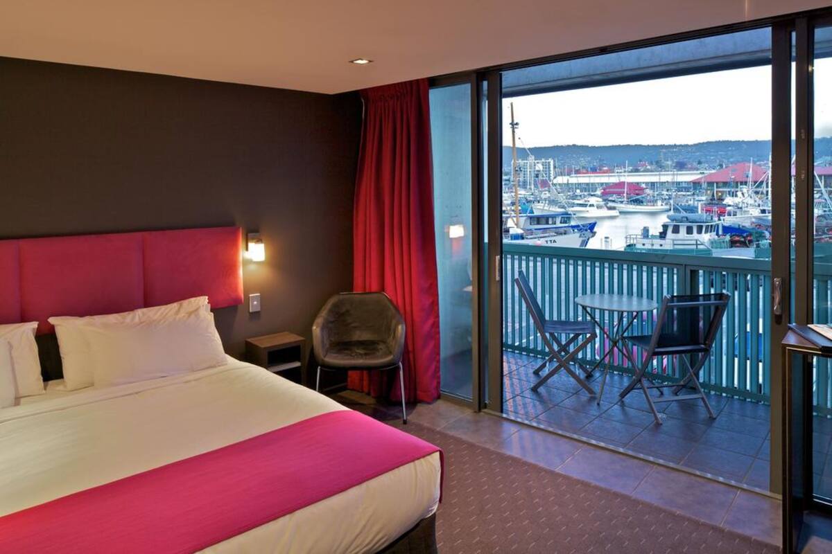 A hotel room with a large bed, a chair, a small table, and a balcony with a view of boats and buildings.