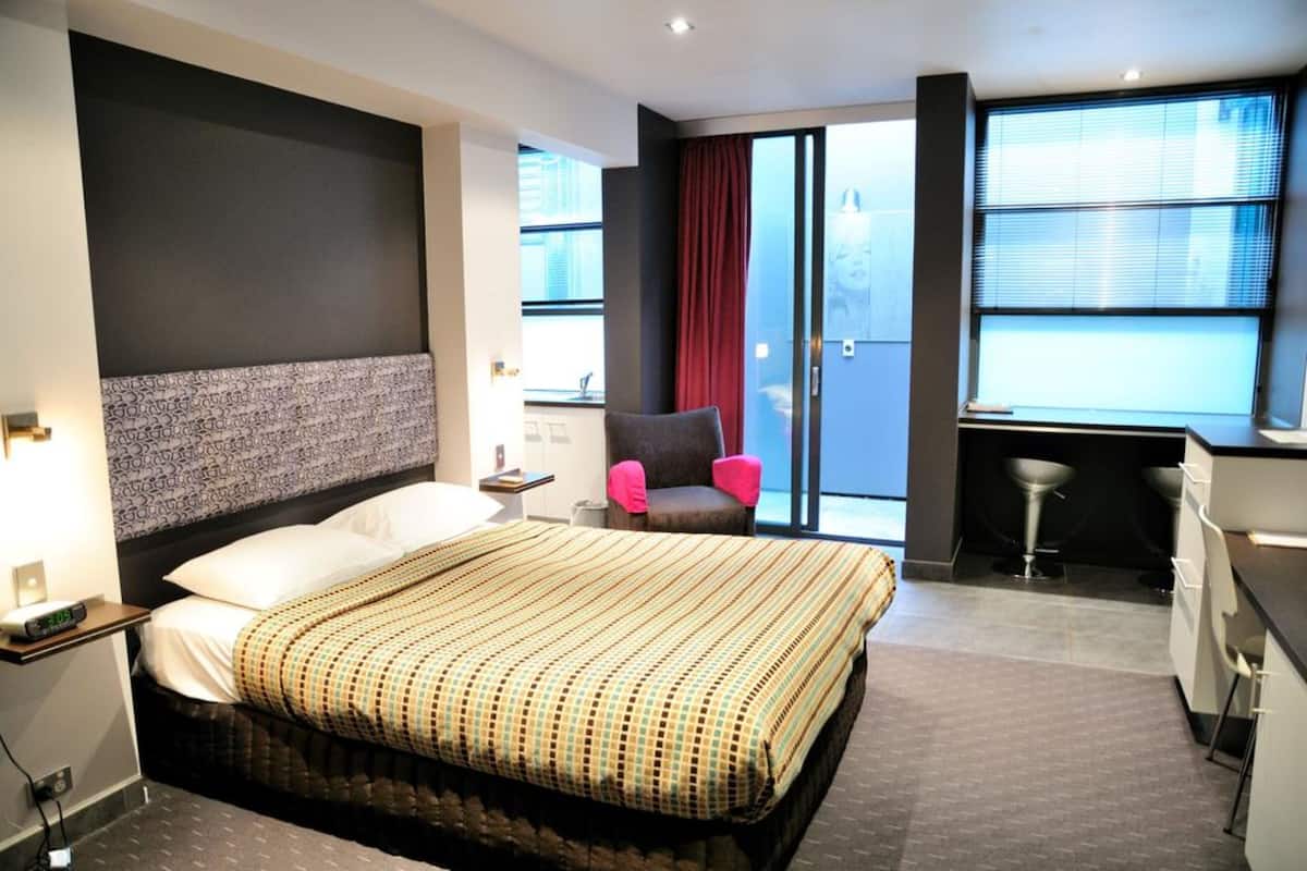 A hotel room with a large bed, a chair, a desk, and a view of the city through large windows.