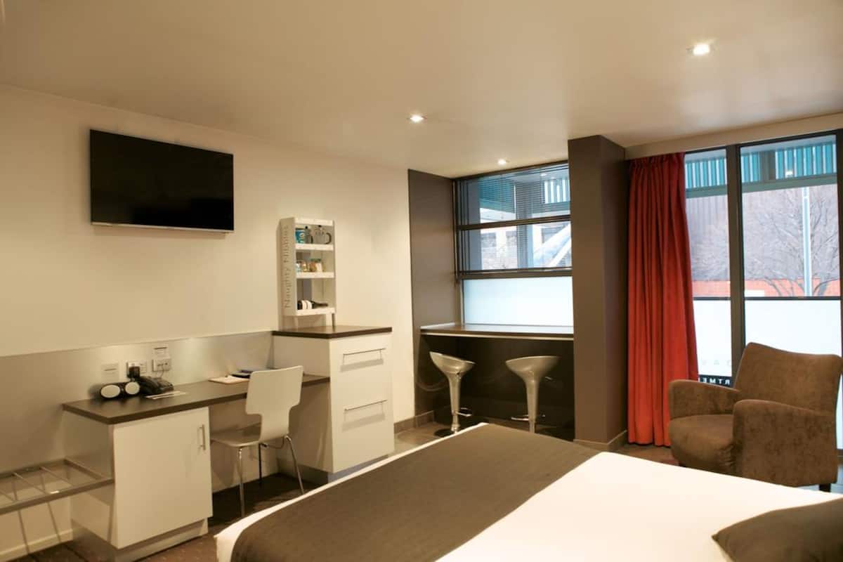 A hotel room with a bed, a desk with a chair, a TV mounted on the wall, a window with blinds, and a red curtain.