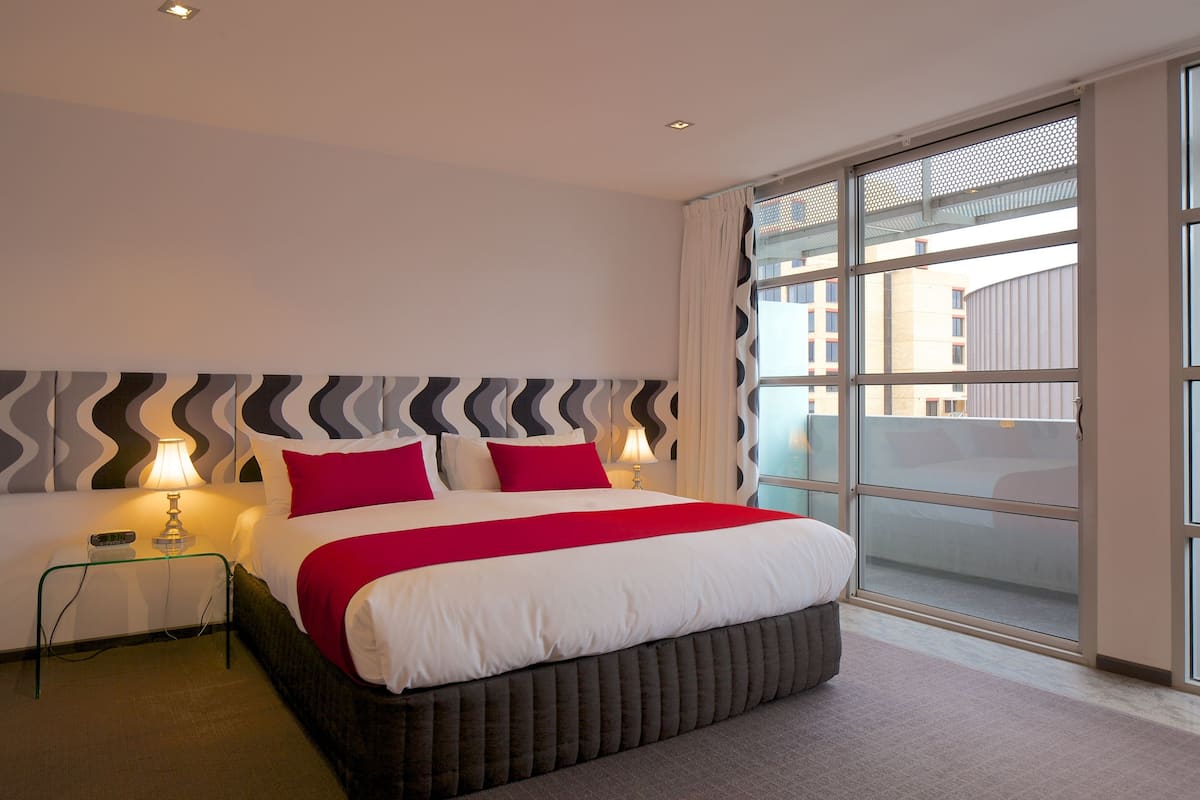 A modern hotel room with a large bed, two bedside lamps, and a view of the city through large windows.
