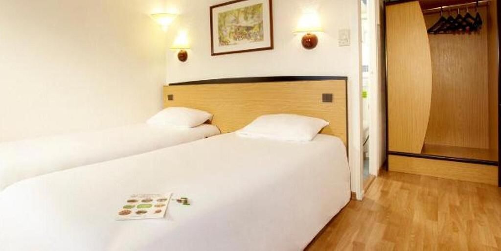 Habitación de hotel con dos camas individuales, cabecero de madera, un cuadro en la pared y un armario de madera con ropa colgada.