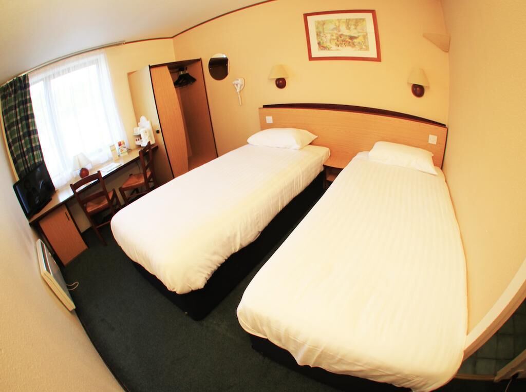 Une chambre d’hôtel avec deux lits simples, un bureau avec une chaise, une petite table et une fenêtre avec des rideaux.