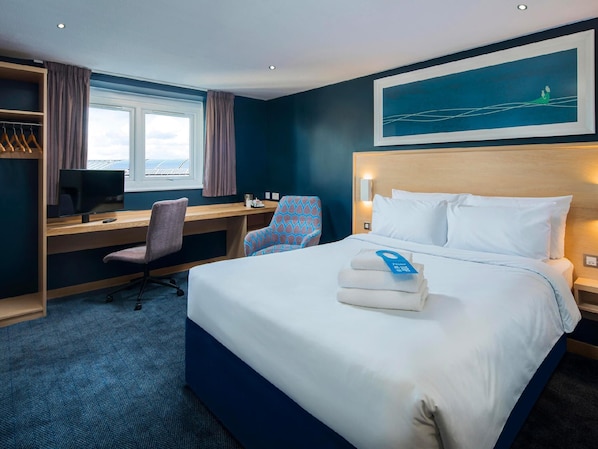 Une chambre d’hôtel avec un lit, un bureau avec un ordinateur, une chaise, une fenêtre et un tableau au mur.