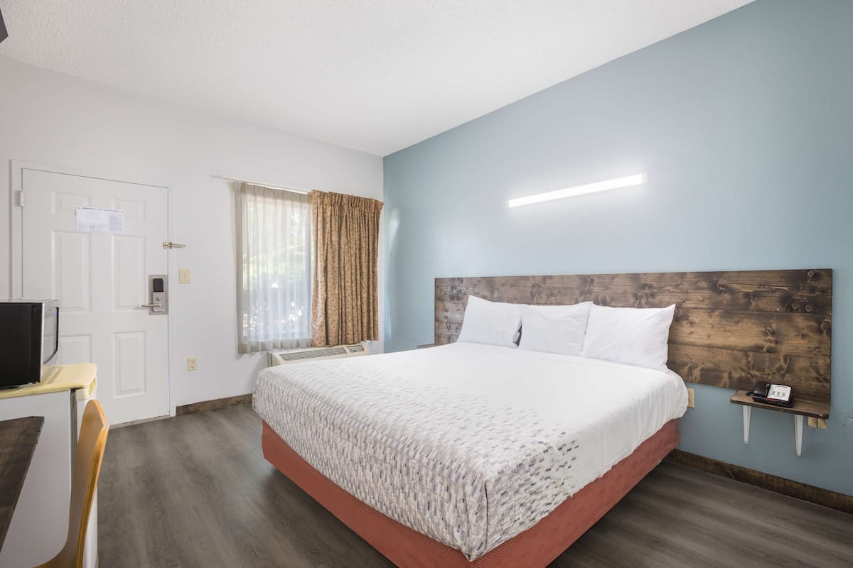 A hotel room with a bed, a desk, a chair, a window with curtains, and a wall-mounted phone.