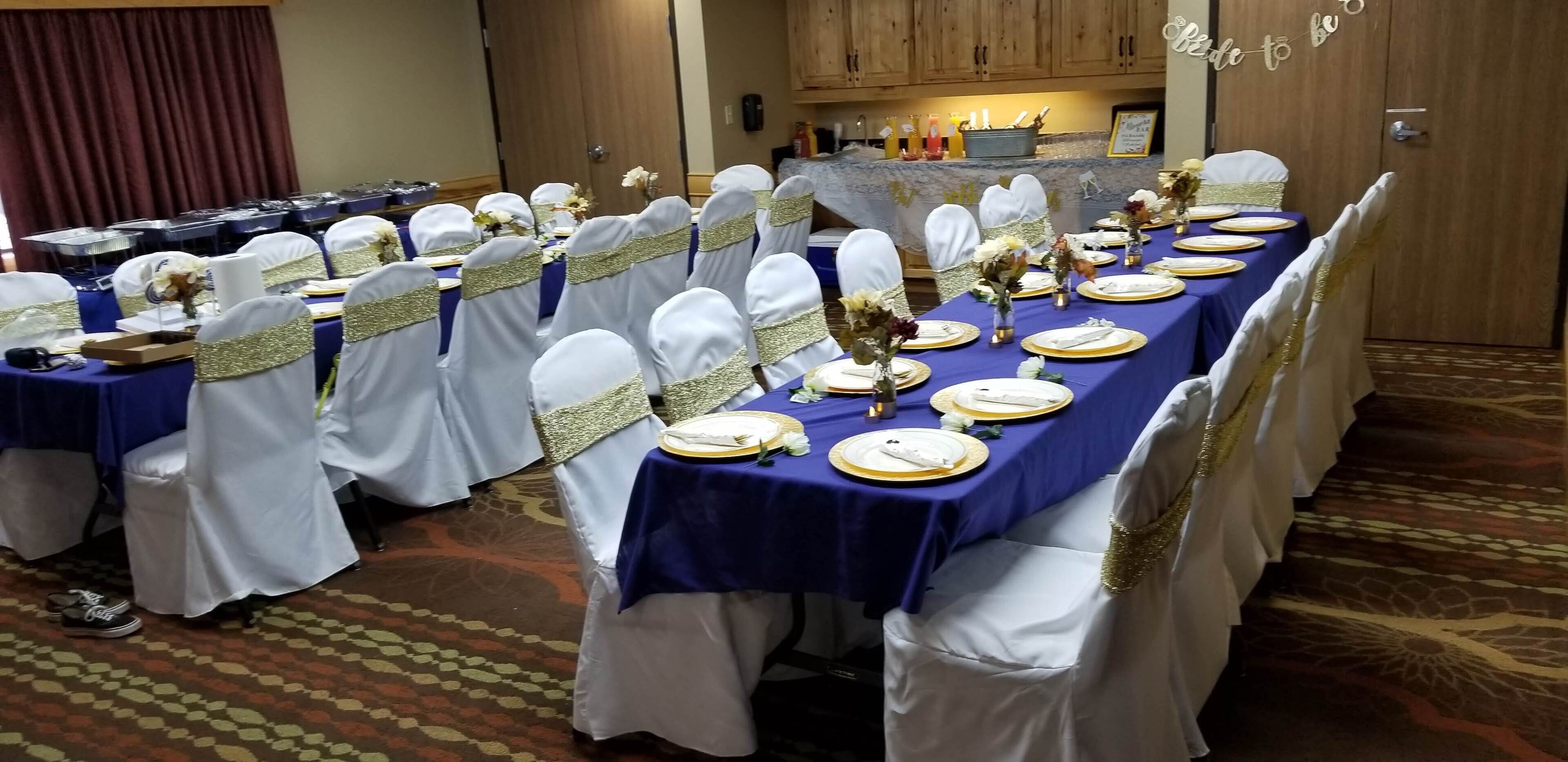 A banquet hall with tables set for a formal event, featuring white chairs with gold sashes, blue tablecloths, and gold-rimmed plates.