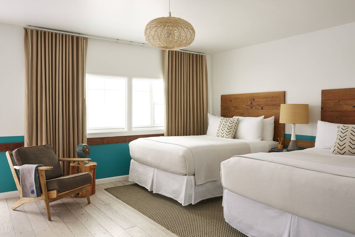 Quarto de hotel com duas camas, uma cadeira de madeira, uma mesinha e um candeeiro de fibra trançada.
