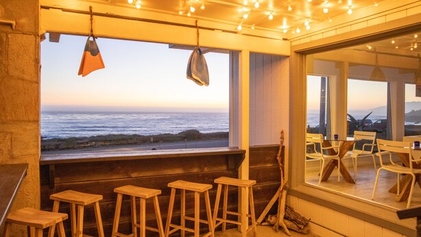 A bar area with wooden stools, a view of the ocean, and hanging lights. - Cambria Beach Lodge (Cambria)