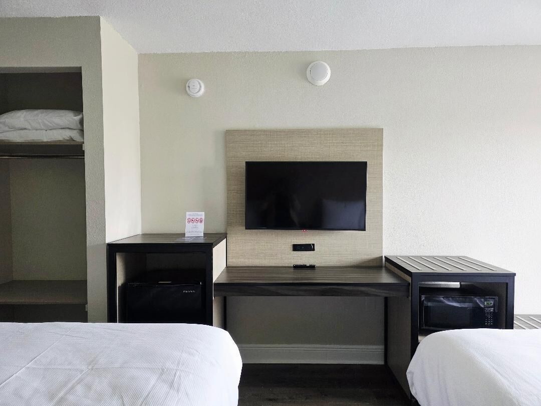 A hotel room with a bed, a desk with a flat-screen TV, a microwave, and a small shelf.