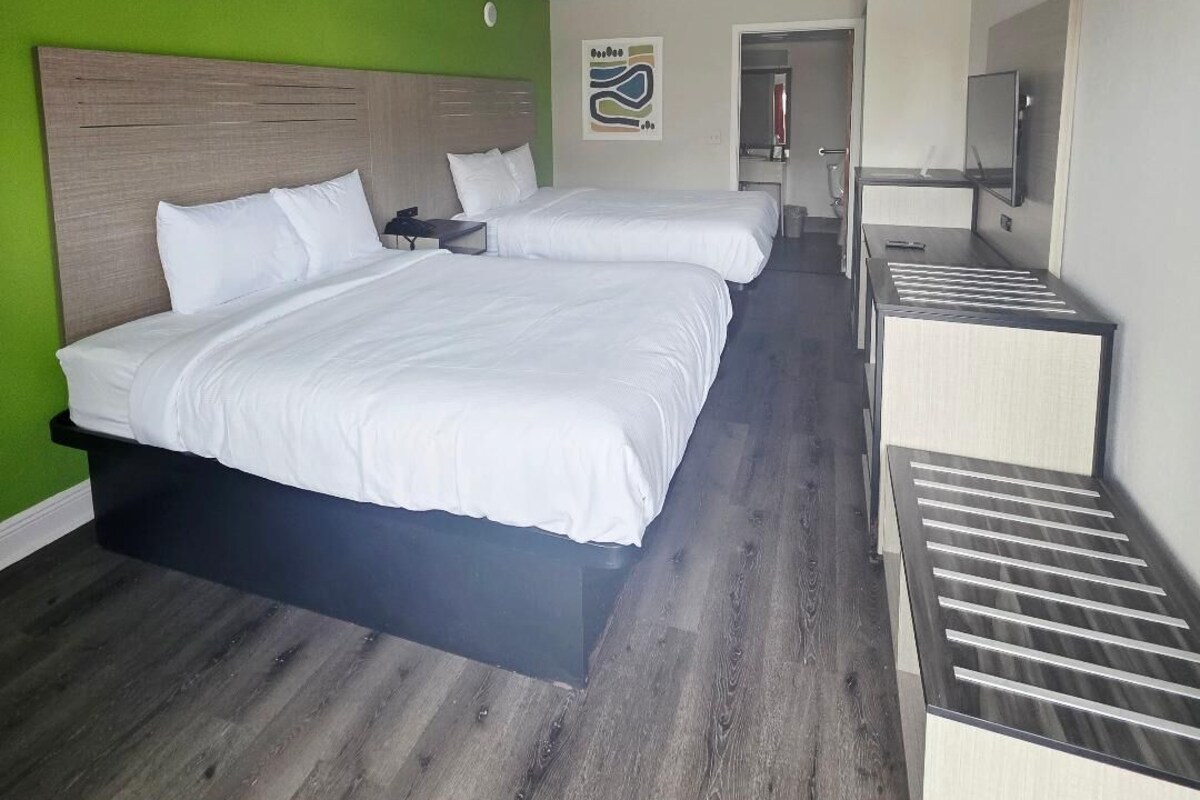 A hotel room with two beds, a wooden floor, a green accent wall, a small desk, and a mirror.