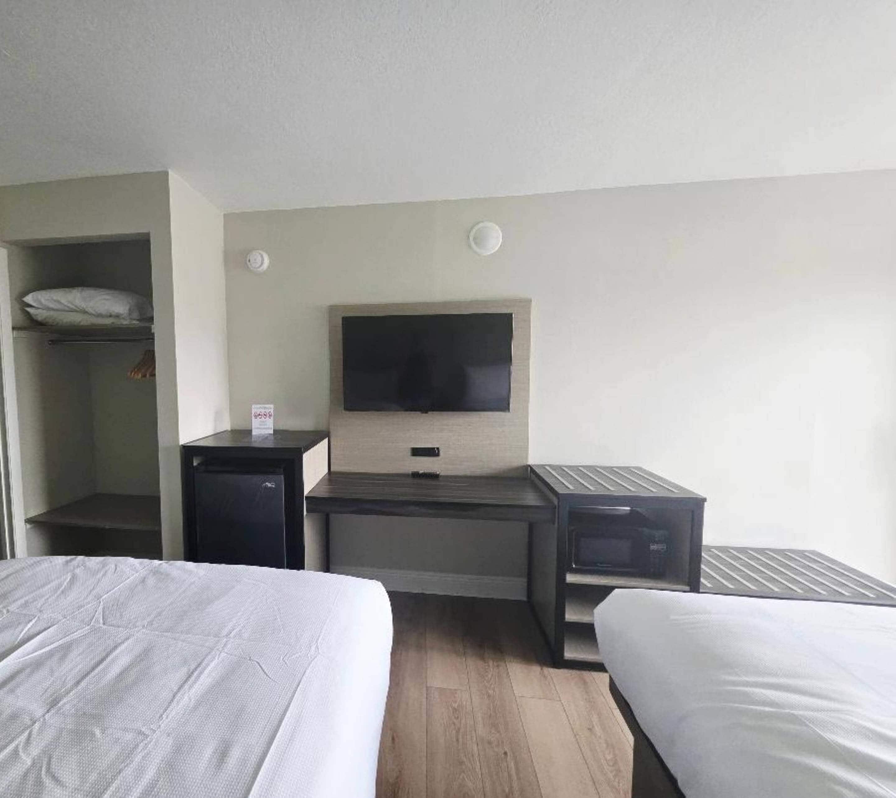 A hotel room with two beds, a desk, a television, a small refrigerator, and a shelf with a pillow.