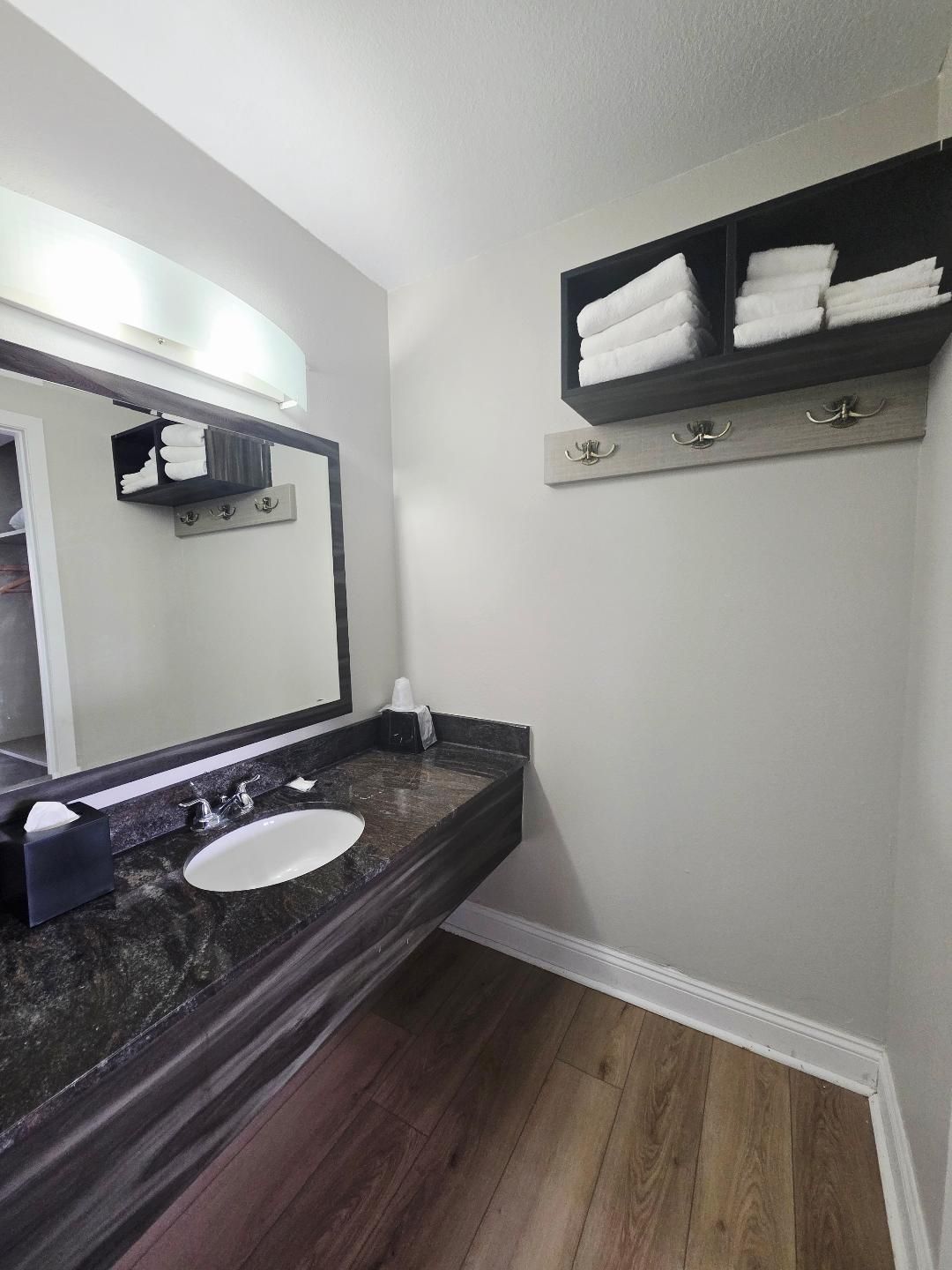 A bathroom with a dark wood floor, a white sink, and a mirror above it. There is a towel rack with towels on the wall.