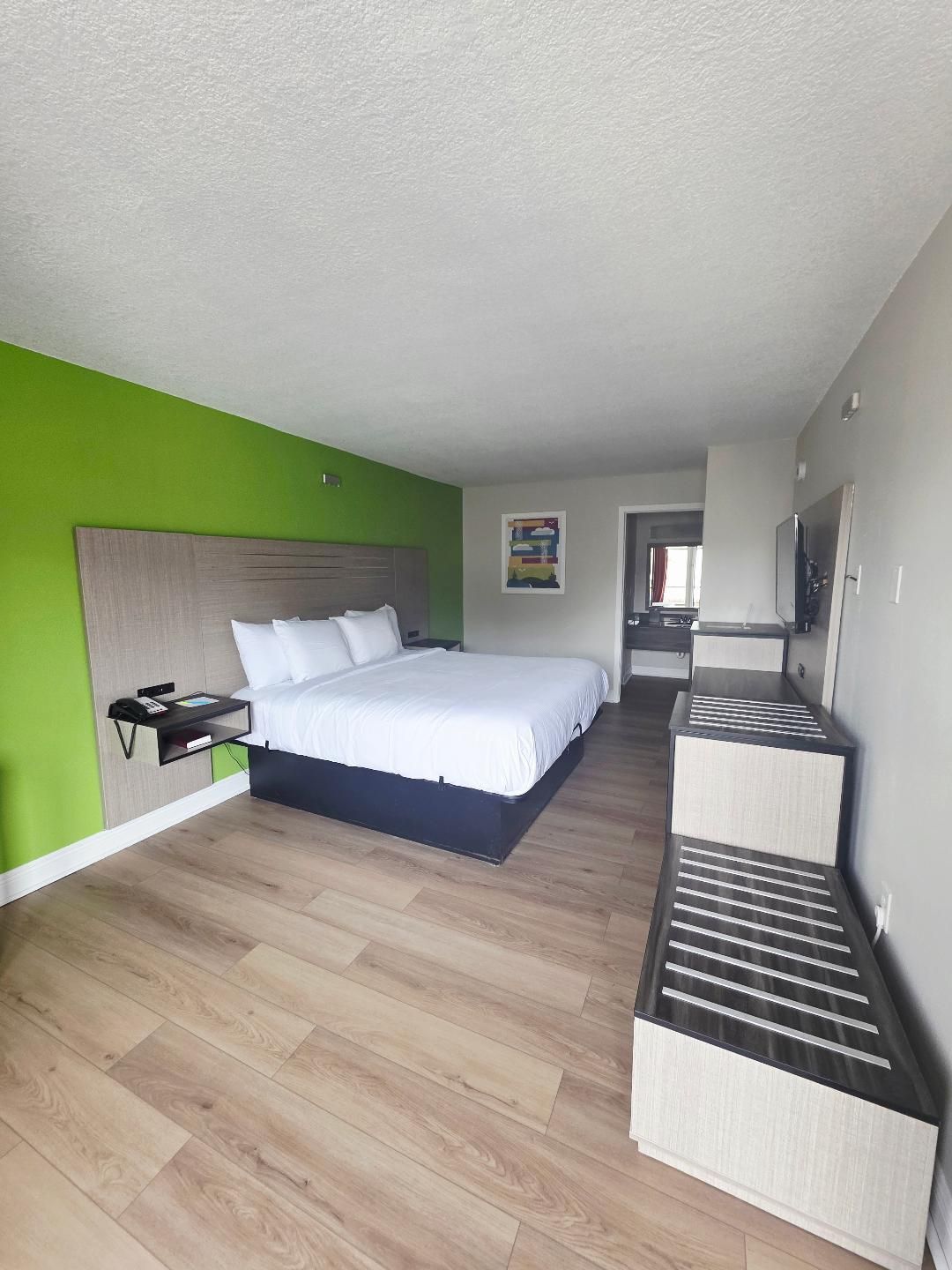 A hotel room with a bed, a bedside table, a TV, and a seating area with a bench.