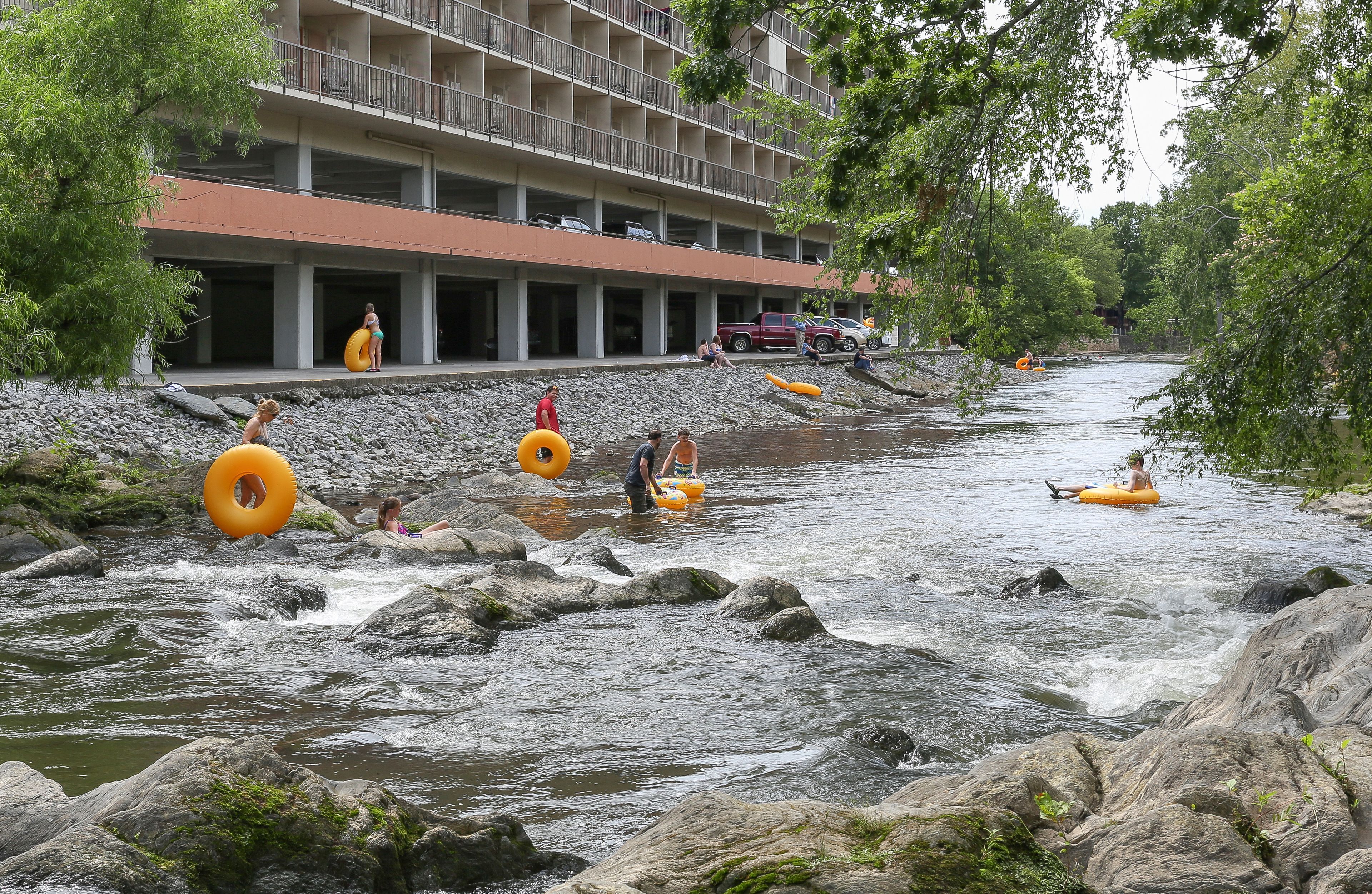 A river with people floating on tubes and rafts. There is a building with balconies and parked cars in the background.