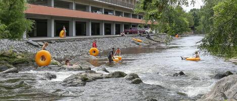 A river with people floating on tubes and rafts. There is a building with balconies and parked cars in the background.