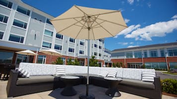 An outdoor seating area with a sofa and two round tables, an umbrella, and a building in the background.