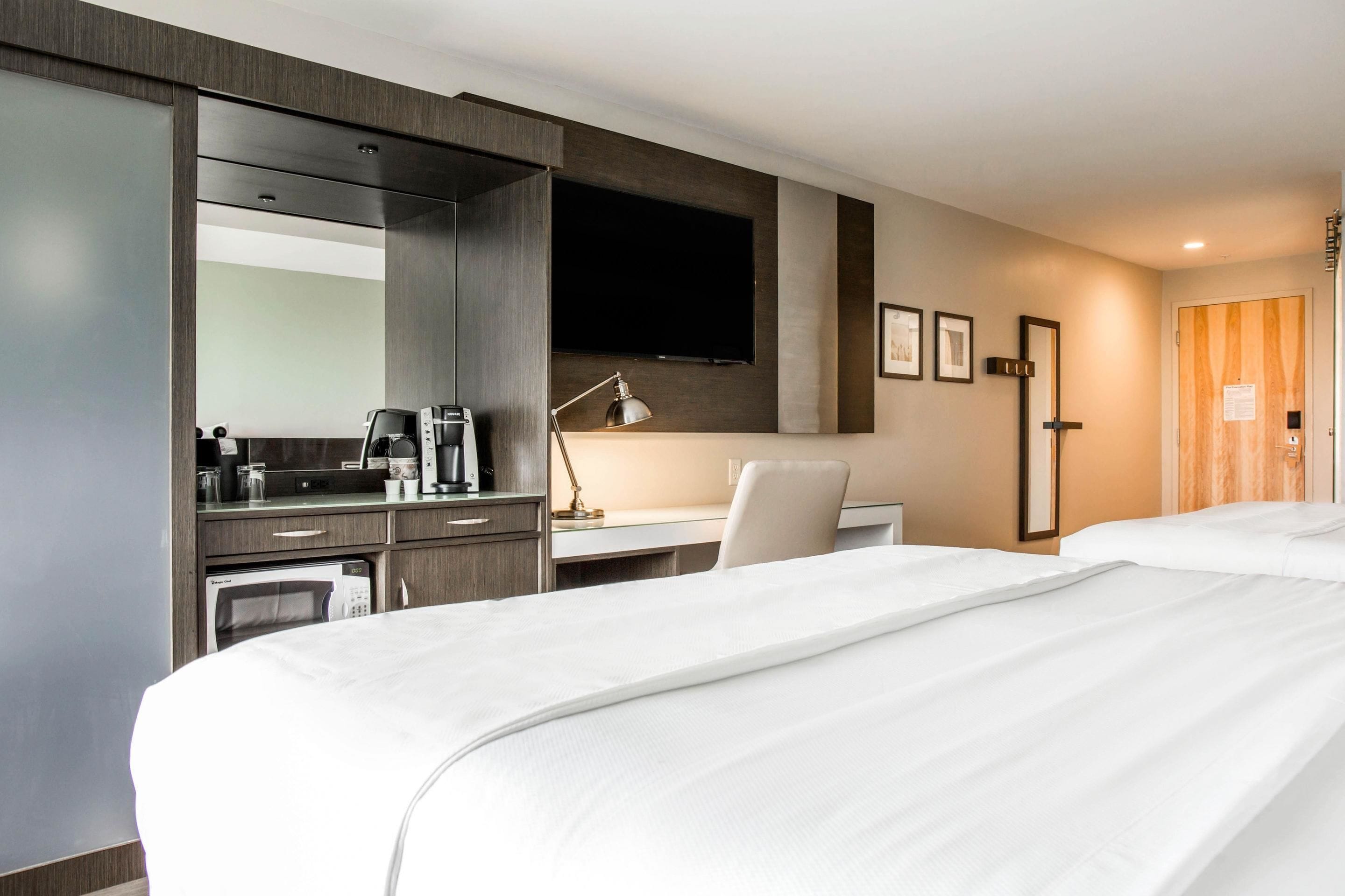 Habitación de hotel con una cama grande, un escritorio con silla, cafetera, televisor de pantalla plana montado en la pared y una puerta con panel de madera.