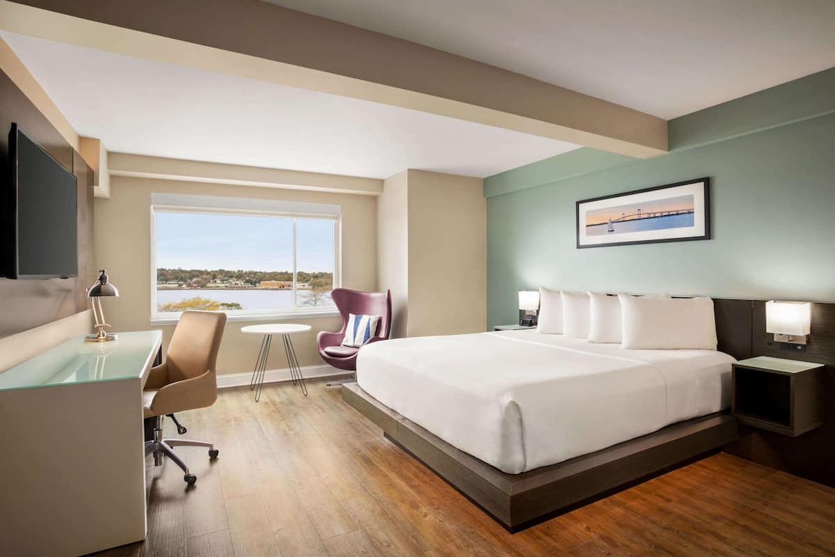 Une chambre d’hôtel avec un grand lit, un bureau, une chaise, une petite table et une fenêtre donnant sur un beau paysage.