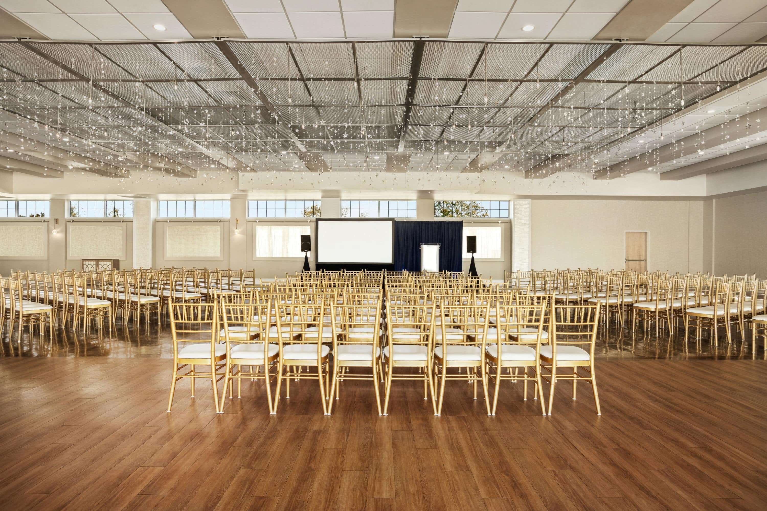 A large hall with rows of chairs arranged for an event, a stage with a screen, and a high ceiling with hanging lights.