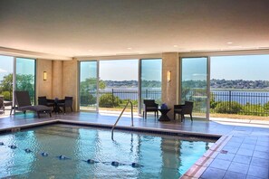 An indoor swimming pool with clear water, surrounded by large windows offering a view of a body of water and greenery.