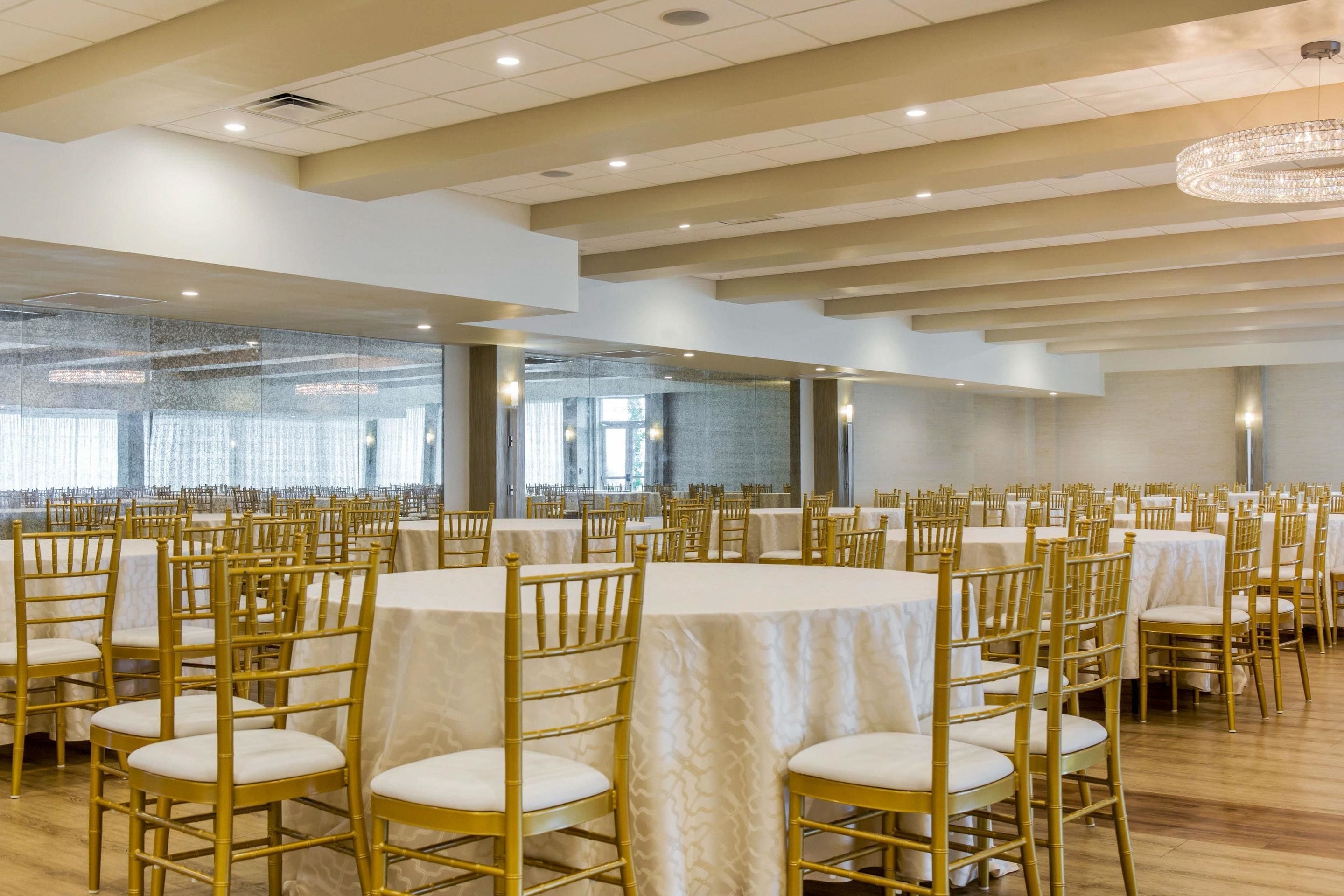 A banquet hall with round tables and gold chairs, large windows, and a chandelier.