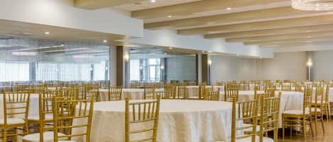 A banquet hall with round tables and gold chairs, large windows, and a chandelier.