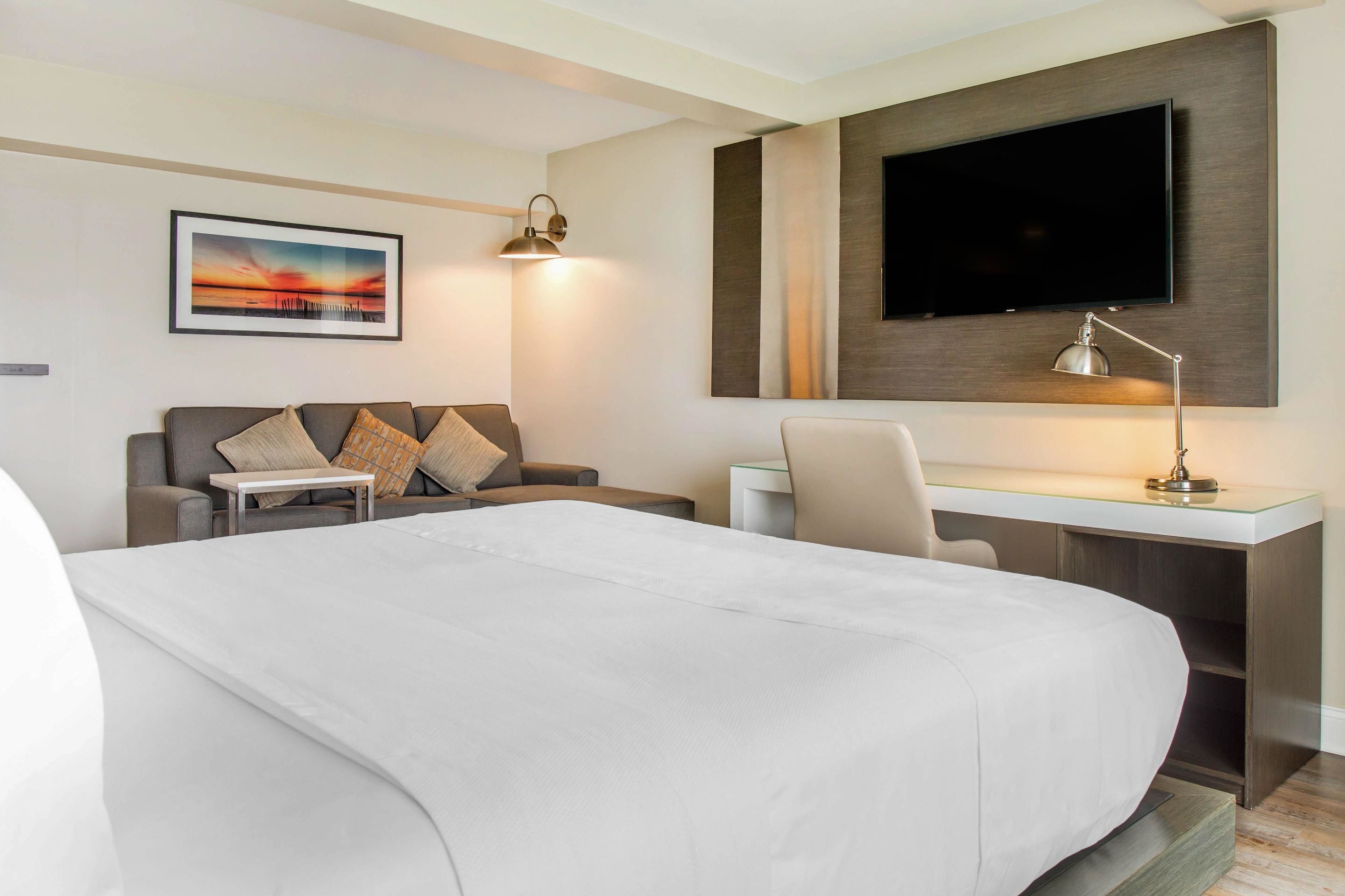 A hotel room with a large bed, a desk with a chair, a television mounted on the wall, and a painting on the wall.