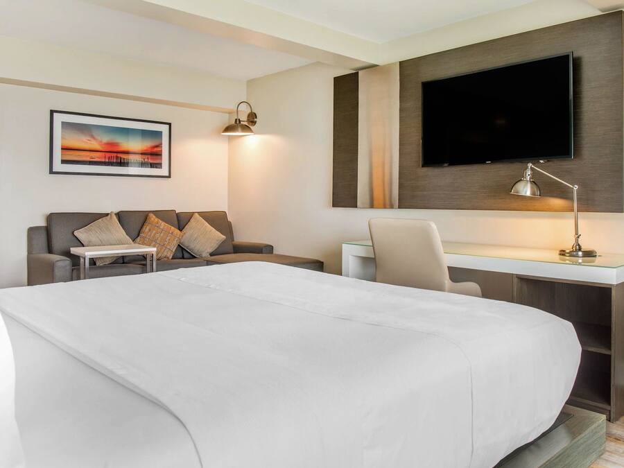 A hotel room with a large bed, a desk with a chair, a television mounted on the wall, and a painting on the wall.