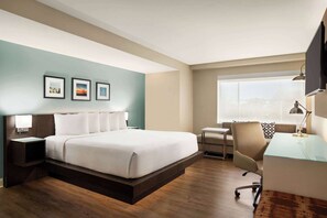 A hotel room with a bed, a desk, a chair, a nightstand, and a window.