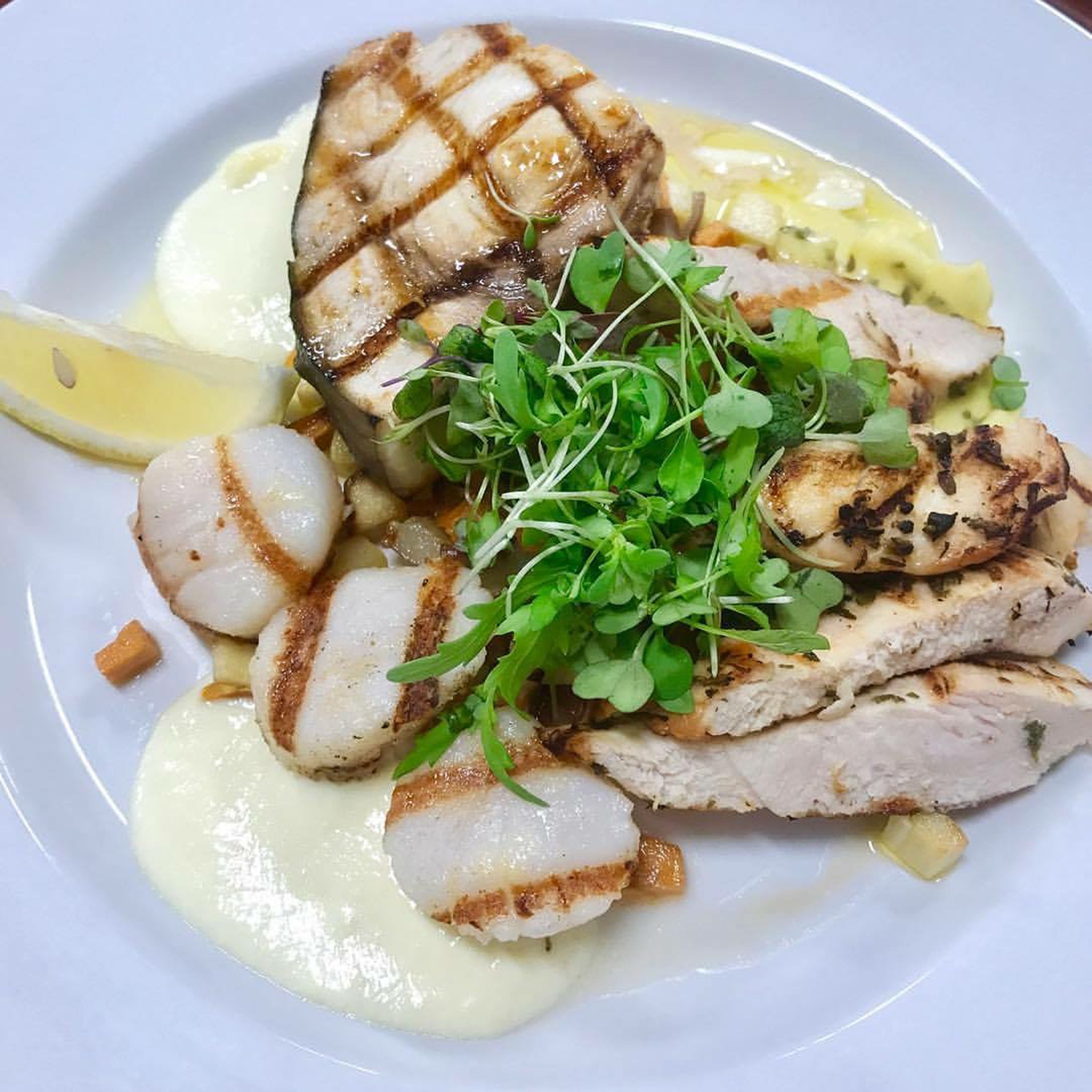 A plate of grilled chicken and scallops with a garnish of greens and a lemon wedge.