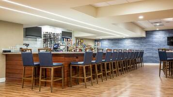 Una zona de bar con una barra alargada, varios taburetes y una estantería llena de botellas de licor.