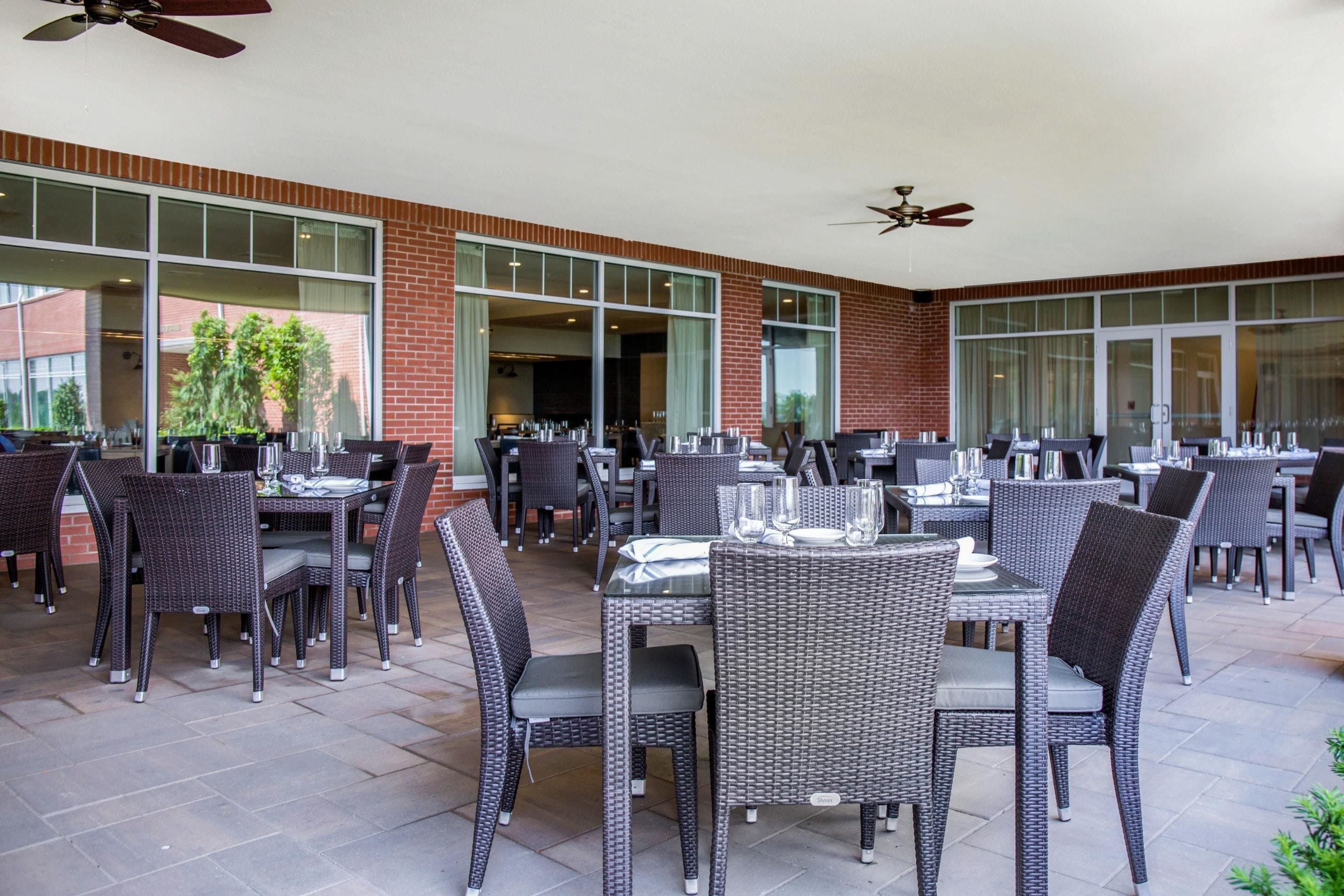 An outdoor patio area with multiple tables and chairs, large windows, and ceiling fans.