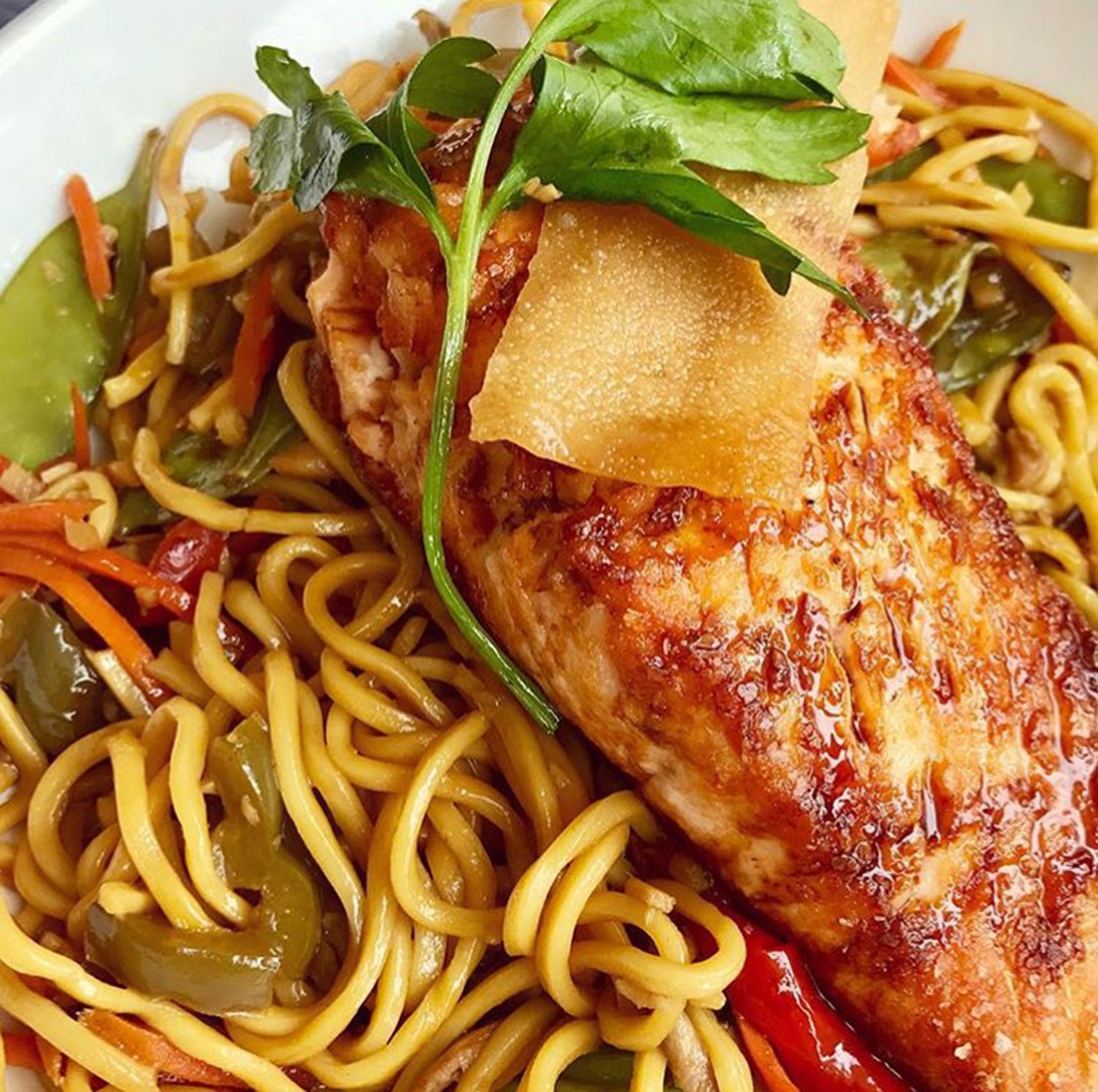 Um prato de noodles refogados com legumes e um pedaço de frango grelhado, coberto com uma folha de manjericão.