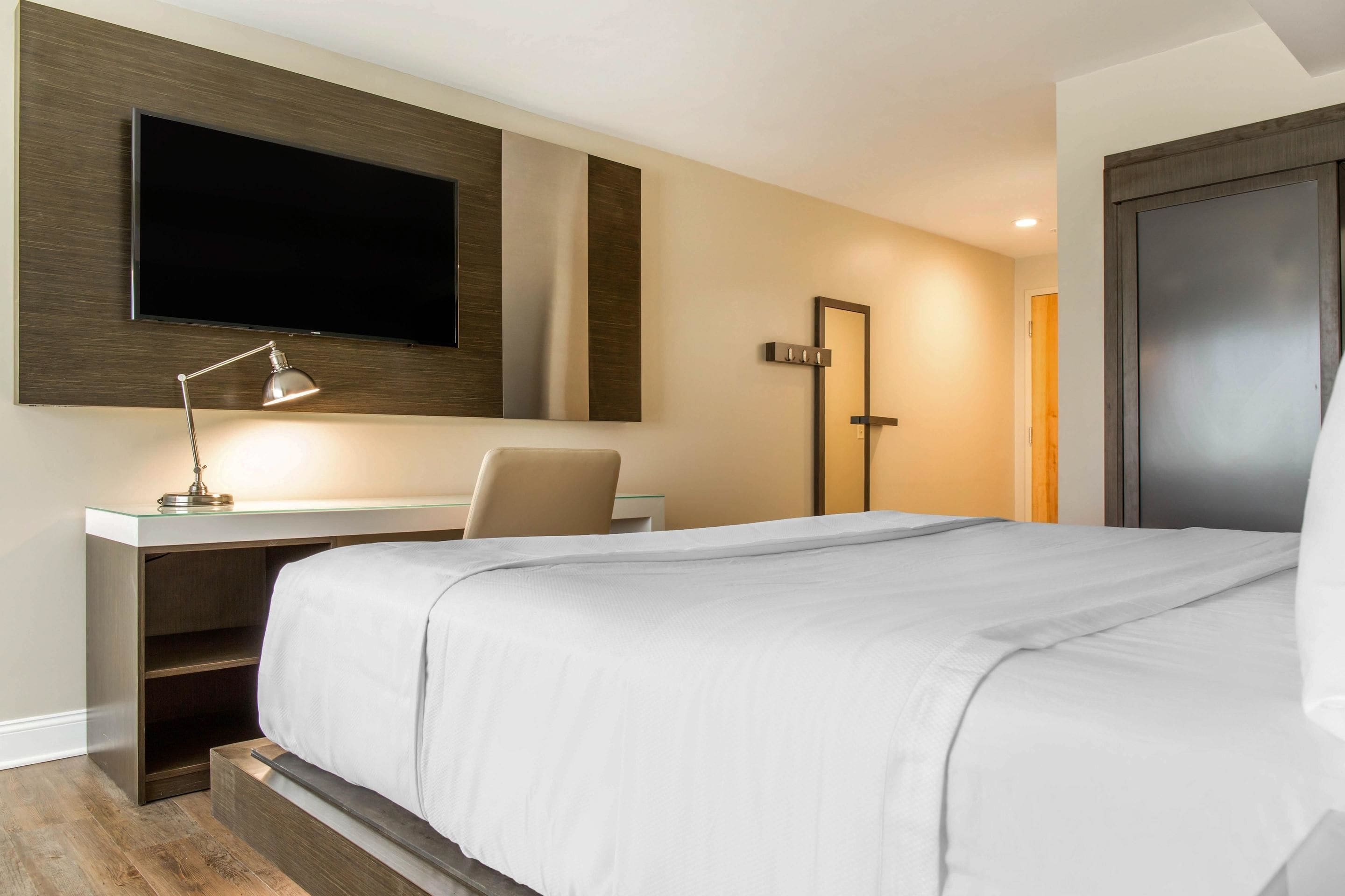 Une chambre d’hôtel moderne dotée d’un grand lit, d’un bureau avec une lampe, d’un téléviseur à écran plat fixé au mur et d’une porte.