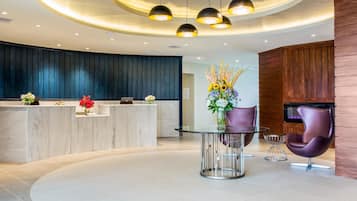A modern reception area with a curved marble counter, a glass table with purple chairs, and a wooden wall with a fireplace.