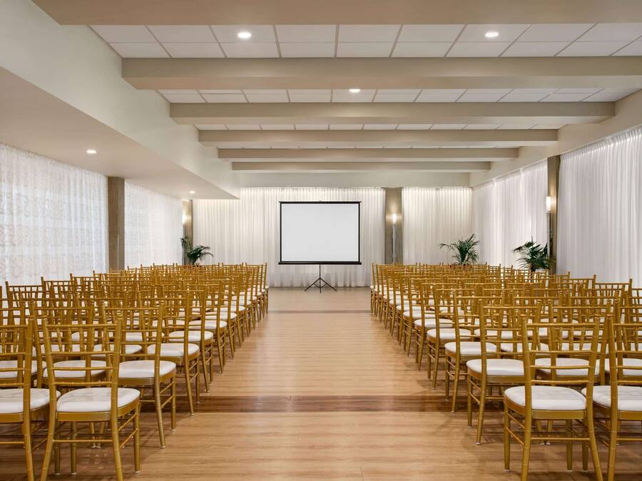 A conference room with rows of golden chairs, a large screen at the front, and white curtains.