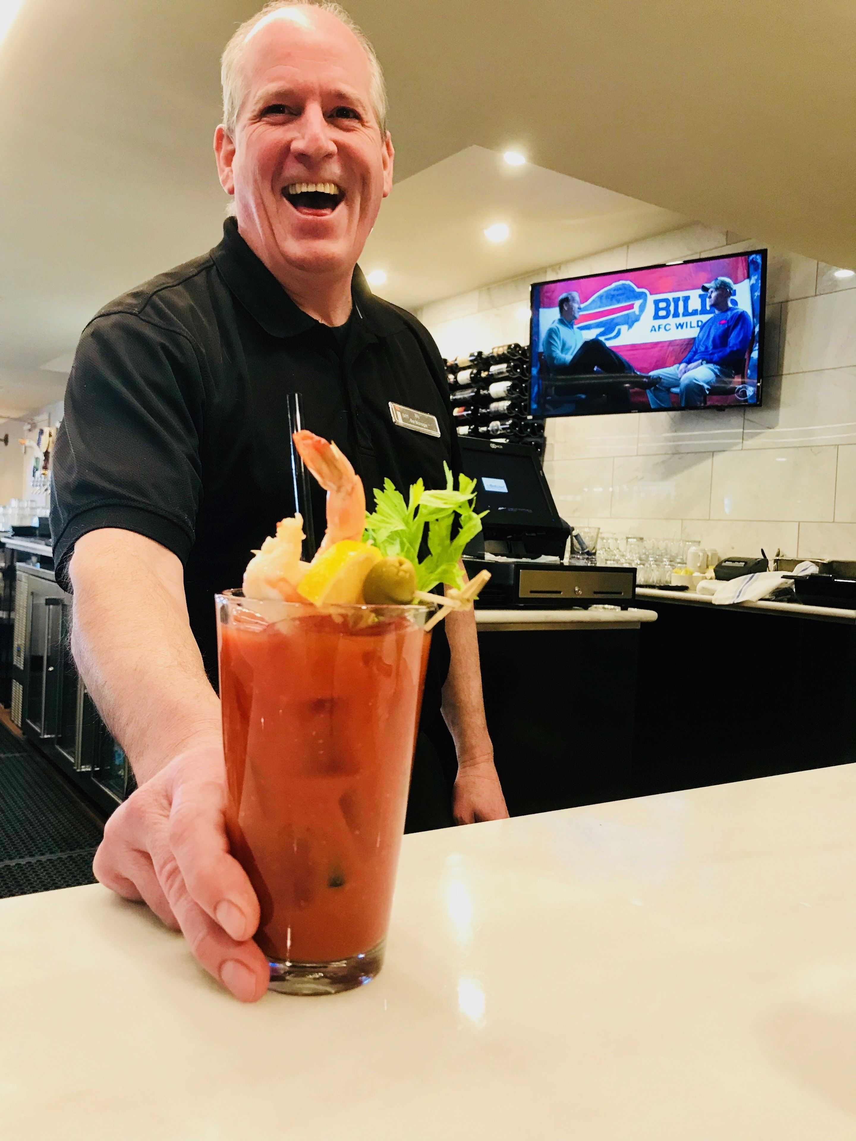 Eine Person hält einen Bloody Mary Cocktail mit Garnitur aus einer Garnele und Sellerie. Im Hintergrund ist ein Fernseher zu sehen, der ein Fußballspiel zeigt.